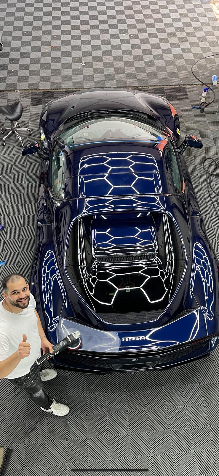 Donkerblauwe Ferrari, van bovenaf gezien in de werkplaats, met de eigenaar ernaast en een polijstmachine in de hand. De hexagonvormige plafondverlichting weerspiegelt zich scherp in het lak.