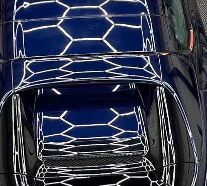Detail van hetzelfde lak: de hexagonvormige lampen weerspiegelen als scherpe witte lijnen in de donkerblauwe motorkap.