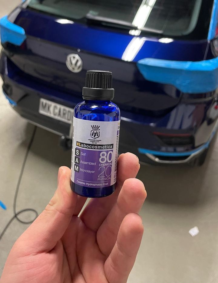 Hand houdt een klein flesje coating vast voor een donkerblauwe auto waarvan de bumper en achterlichten met blauwe tape zijn afgeplakt.