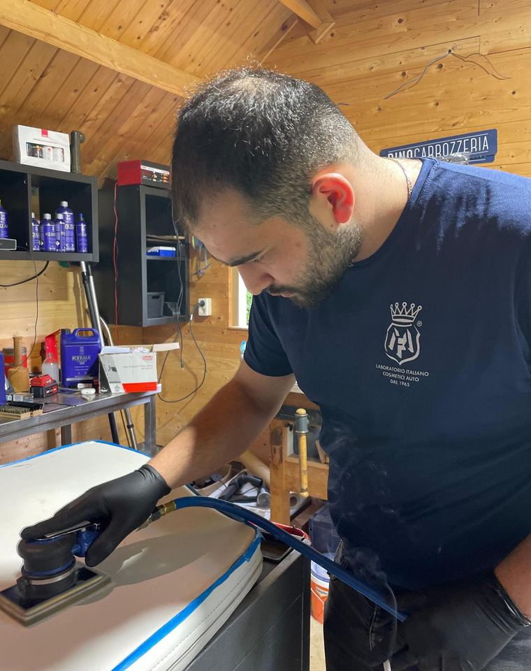 Man met zwarte handschoenen polijst met een machine een wit paneel dat langs de randen met blauwe tape is afgeplakt.