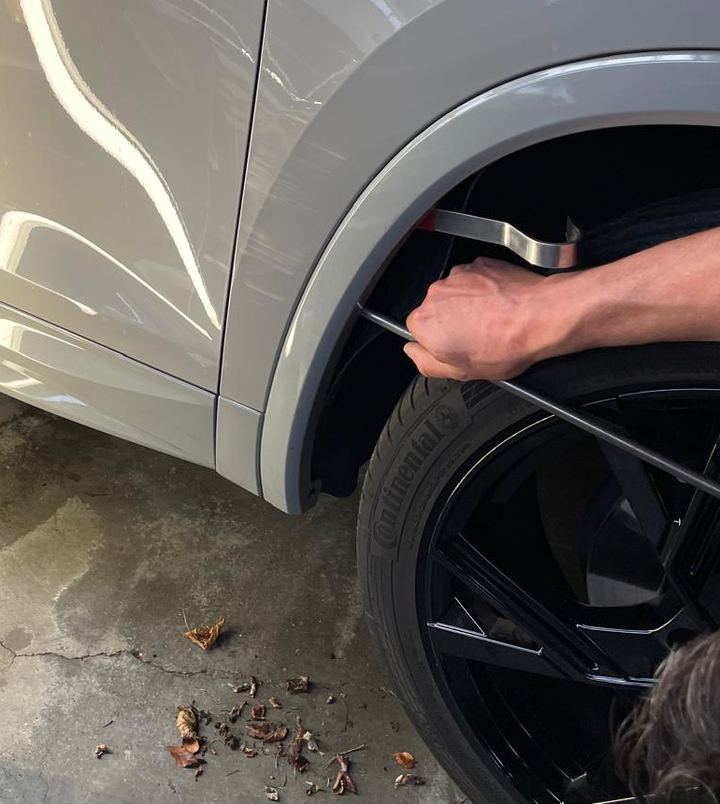 Hand met een lange uitdeukhaak die via de wielkast achter het plaatwerk van een grijze auto reikt.